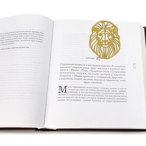 May include: A gold-coloured metal bookmark shaped like a lion's head. The bookmark is on a page of a book with text in Russian. The text on the page reads "лекция 27" and "Сохранение припасов в жестяных ящиках".