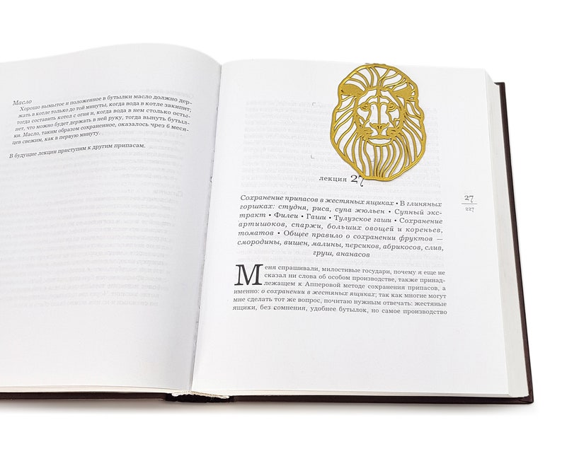 May include: A gold-coloured metal bookmark shaped like a lion's head. The bookmark is on a page of a book with text in Russian. The text on the page reads "лекция 27" and "Сохранение припасов в жестяных ящиках".