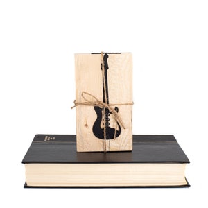 Black metal electric guitar bookmark tied to a wooden block printed with its silhouette and the phrase Reading is good for you, displayed on a closed book. A cool gift for music-loving readers.