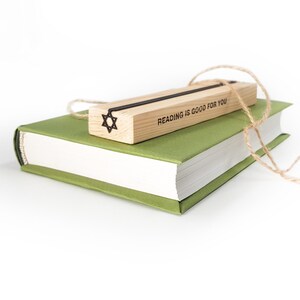 Black Star of David metal bookmark resting on a wooden gift block engraved with “Reading is good for you,” placed atop a green hardcover book with rustic twine. Made by Coolbookmark on Etsy. Not mass-manufactured.