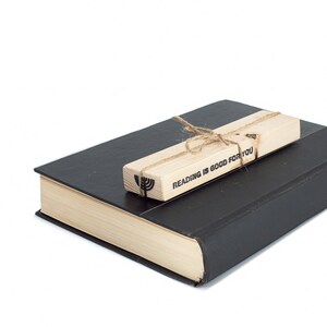 Black metal menorah bookmark tied to a rustic wooden block printed with ‘Reading is good for you,’ resting on a closed book. A meaningful handmade Judaica gift.