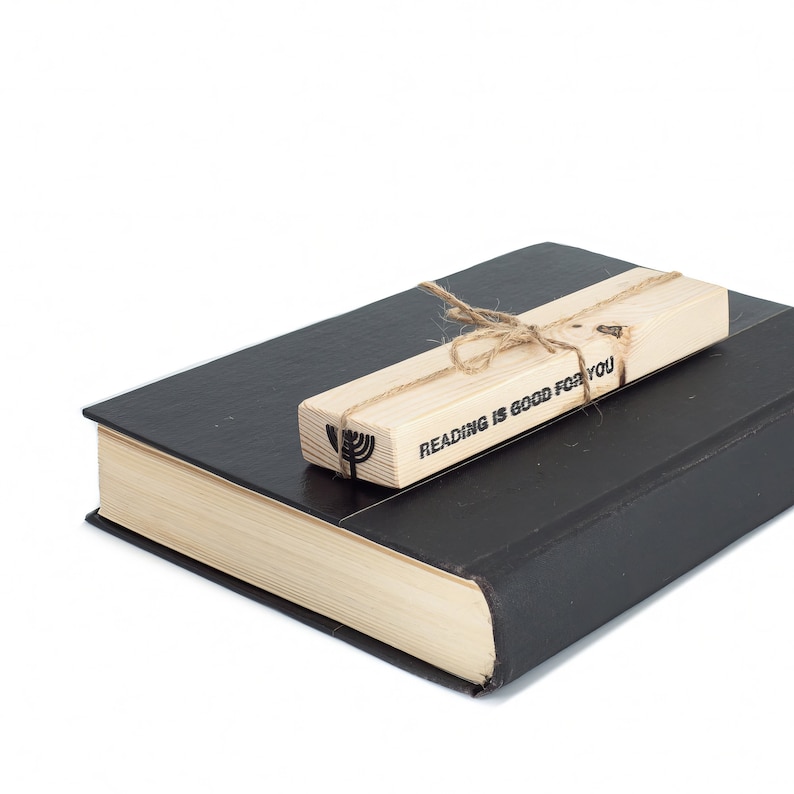 Black metal menorah bookmark tied to a rustic wooden block printed with ‘Reading is good for you,’ resting on a closed book. A meaningful handmade Judaica gift.
