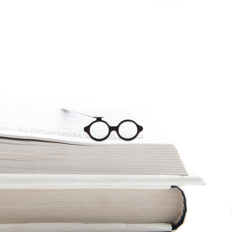 Black metal reading accessory shaped like round architect glasses, resting on the edge of a stacked open book. Clean, Bauhaus-inspired design.