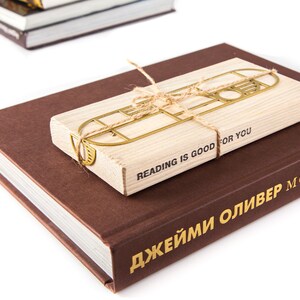 Gold metal bookmark Bauhaus Geometry attached to a wooden packaging block. The bookmark is tied with twine to it,  the text 'Reading is good for you is printed on it. The block is sitting on top of a brown book with the text 'ДЖЕЙМИ ОЛИВЕР' printed on the spine.
