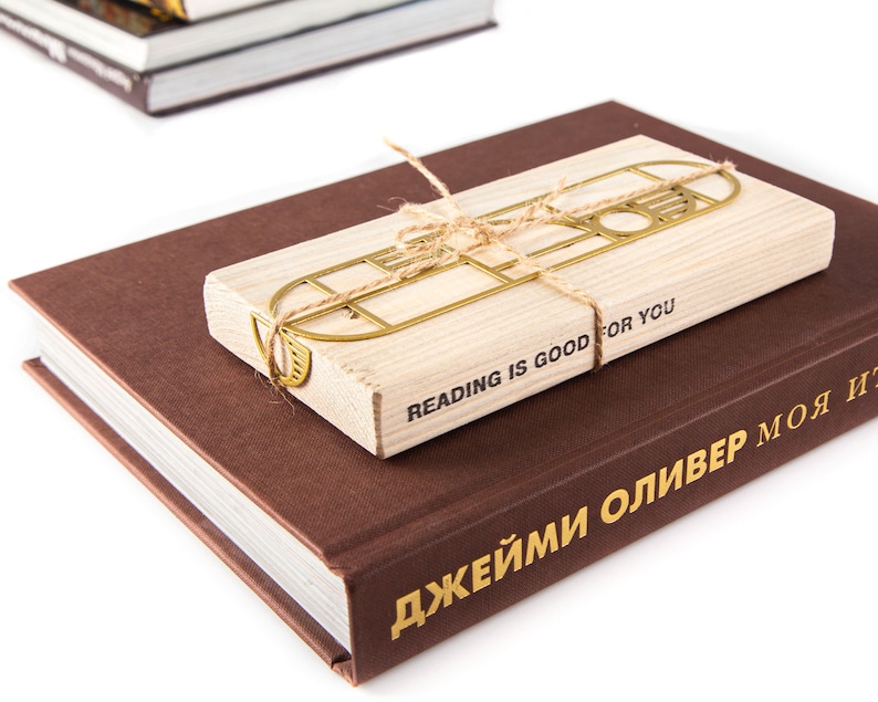 Gold metal bookmark Bauhaus Geometry attached to a wooden packaging block. The bookmark is tied with twine to it,  the text 'Reading is good for you is printed on it. The block is sitting on top of a brown book with the text 'ДЖЕЙМИ ОЛИВЕР' printed on the spine.