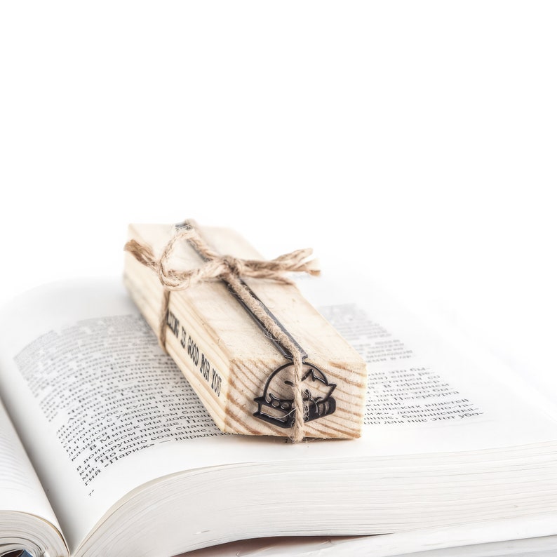 Op de afbeelding: Een houten boekenlegger met een kat- en boekontwerp, vastgebonden met touw, rust op een open boek. De boekenlegger heeft de tekst "HOW IS GOOD FOR YOU" erop gedrukt. De boekenlegger is gemaakt van lichtgekleurd hout.