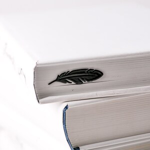 May include: Two white books stacked on top of each other. The top book has a black feather bookmark on the spine.