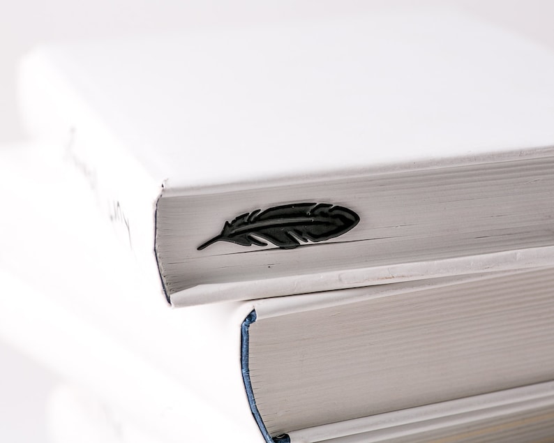 May include: Two white books stacked on top of each other. The top book has a black feather bookmark on the spine.