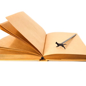 Black metal running cat bookmark placed on the open pages of a blank kraft paper notebook.