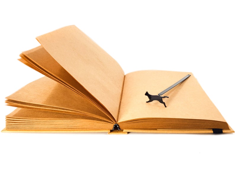 Black metal running cat bookmark placed on the open pages of a blank kraft paper notebook.