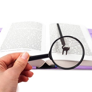 May include: An open book with a purple cover and a black cat bookmark. A magnifying glass is positioned over the text, enhancing readability. The image is set against a white background, highlighting the book and reading accessories.