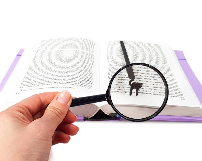 May include: An open book with a purple cover and a black cat bookmark. A magnifying glass is positioned over the text, enhancing readability. The image is set against a white background, highlighting the book and reading accessories.