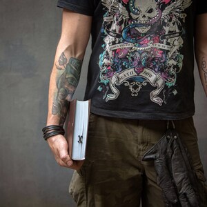 Może przedstawiać: Osoba w czarnym t-shircie z grafiką czaszki i węża, z tekstem "Death Row" i "Fearless", trzymająca czarną książkę ze srebrnym zapięciem i srebrnym "X" na okładce. Osoba ma tatuaże na obu ramionach.