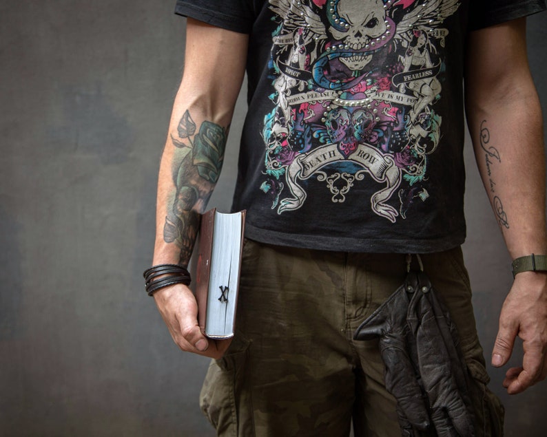 Może przedstawiać: Osoba w czarnym t-shircie z grafiką czaszki i węża, z tekstem "Death Row" i "Fearless", trzymająca czarną książkę ze srebrnym zapięciem i srebrnym "X" na okładce. Osoba ma tatuaże na obu ramionach.