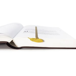 May include: A gold metal feather bookmark is placed in a book with white pages. The book is open and the bookmark is sticking out of the top of the page.