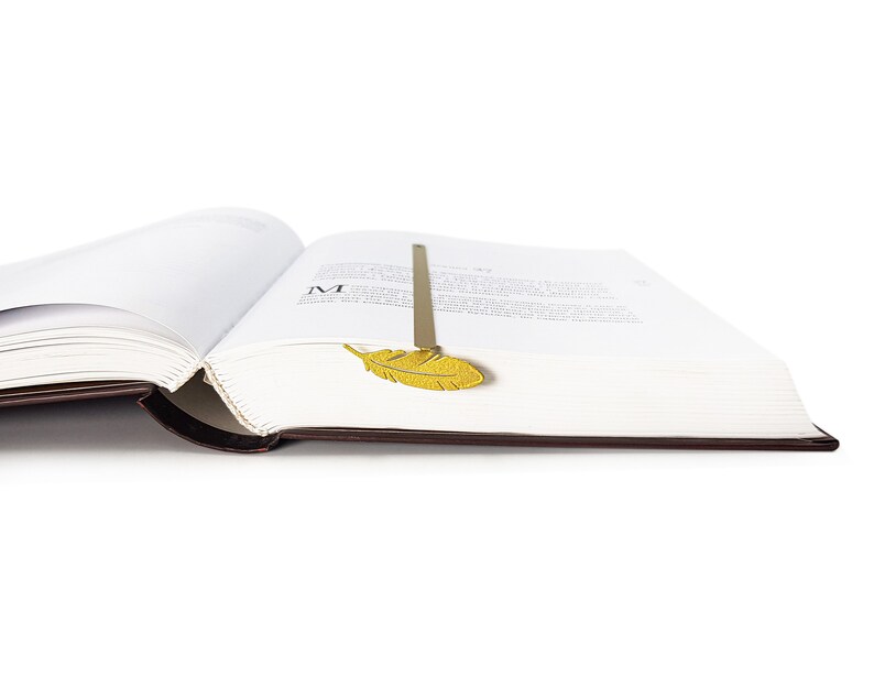 May include: A gold metal feather bookmark is placed in a book with white pages. The book is open and the bookmark is sticking out of the top of the page.