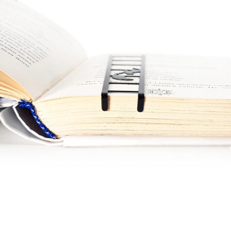 Black metal cat bookmark inserted between book pages, ladder design sitting flat and visible at the top of a thick open book.