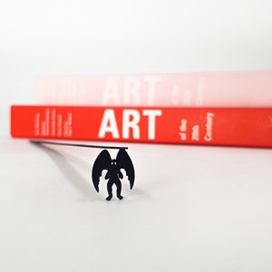 Op de afbeelding: Een zwart metalen boekenlegger met een silhouet van een gevleugelde demon. De boekenlegger is geplaatst voor twee boeken, een rode en een roze, met het woord "ART" zichtbaar op de rug van het rode boek.