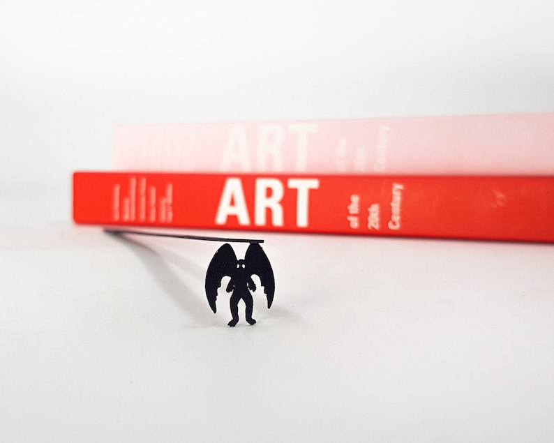 Black metal laser cut cryptid bookmark Mothman. A long flat metal stick is sitting between the pages of the book. The mothman is seen on the side of the book even when the book is closed.