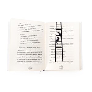 Black metal cat bookmark shaped like a ladder placed vertically between open book pages, with two cats climbing and visible above the text.