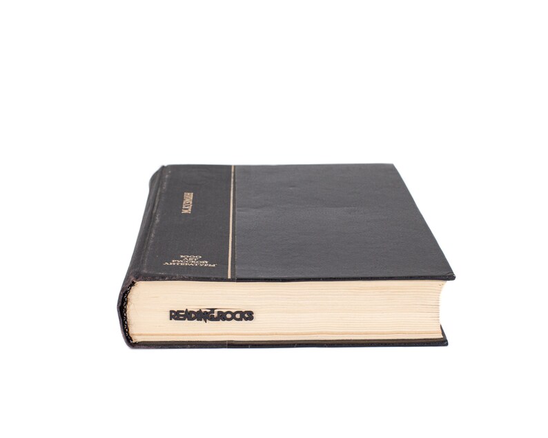Black metal page saver with the phrase ‘READING ROCKS’ peeking from a closed hardcover book. A fun and bold gift for book lovers.