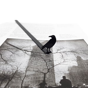 Black metal page saver shaped like a raven, perched on a grayscale photo of a foggy cityscape. A dark, poetic accessory for lovers of gothic fiction and urban mystery.