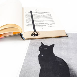 Op de afbeelding: Een zwartmetalen boekenlegger met een kat die op een boek slaapt, geplaatst in een open boek. De boekenlegger bevindt zich boven een zwart-wit afbeelding van een zwarte kat. Het boek heeft verouderde, vergeelde pagina's.