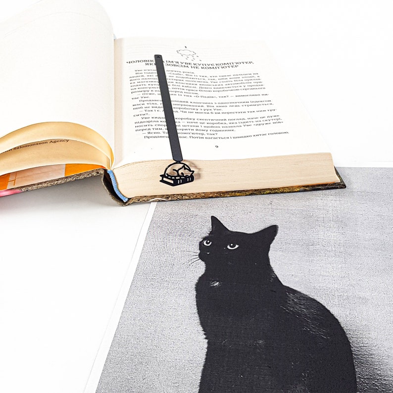 Op de afbeelding: Een zwartmetalen boekenlegger met een kat die op een boek slaapt, geplaatst in een open boek. De boekenlegger bevindt zich boven een zwart-wit afbeelding van een zwarte kat. Het boek heeft verouderde, vergeelde pagina's.