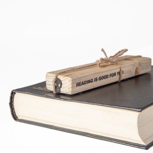 May include: A wooden bookmark with the text "Reading is good for you" tied with twine. The bookmark is resting on top of a closed black hardcover book.