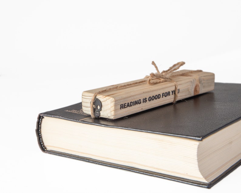May include: A wooden bookmark with the text "Reading is good for you" tied with twine. The bookmark is resting on top of a closed black hardcover book.