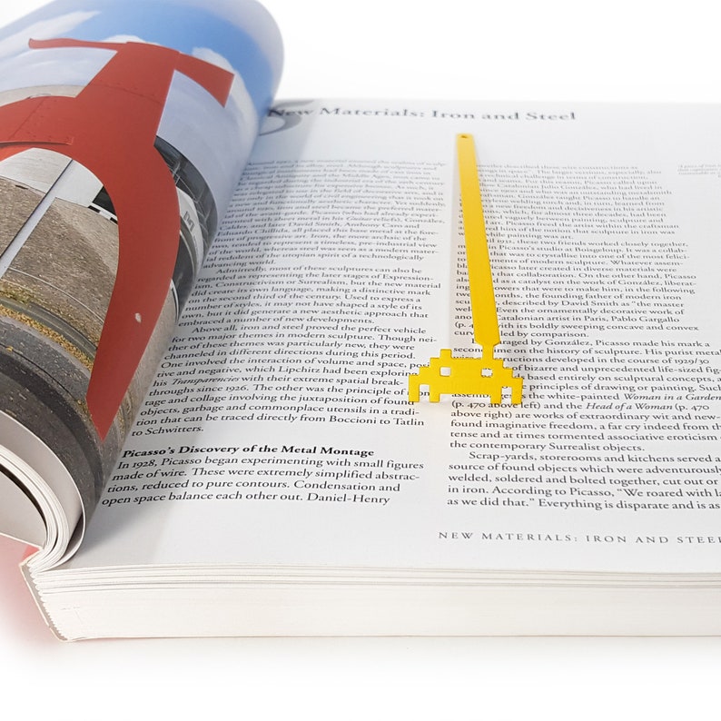 May include: A yellow metal bookmark shaped like a spaceship with a book open to a page about the history of sculpture. The text on the page discusses the use of iron and steel in modern sculpture.