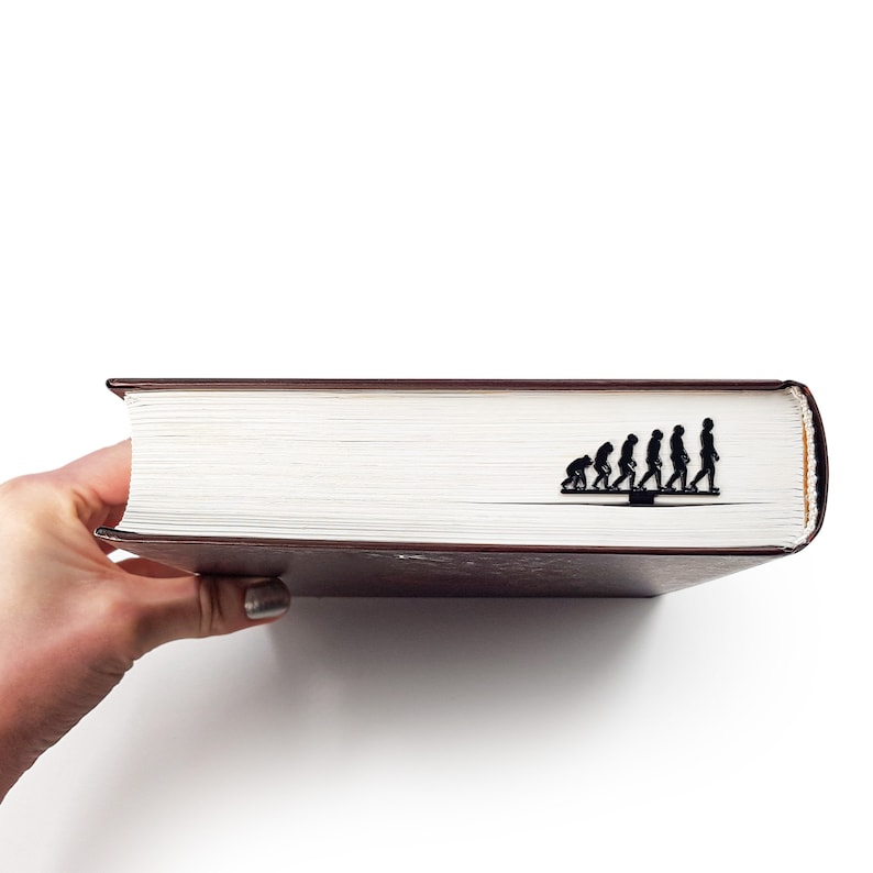 May include: A black bookmark shaped like a line of stick figures depicting the evolution of humans. The bookmark is on top of a closed brown book.