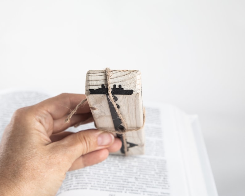 May include: A wooden bookmark with a black ship silhouette design. The bookmark is tied with twine and is being held by a hand.