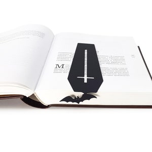 Black metal coffin-shaped bookmark with a cross cutout and bat detail placed inside an open book, with the bat peeking out from the bottom edge of the pages.