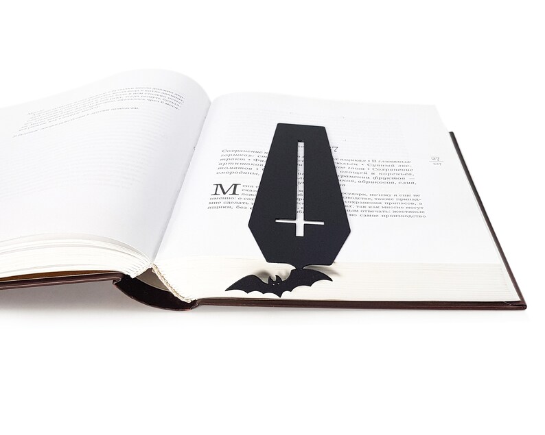 Black metal coffin-shaped bookmark with a cross cutout and bat detail placed inside an open book, with the bat peeking out from the bottom edge of the pages.