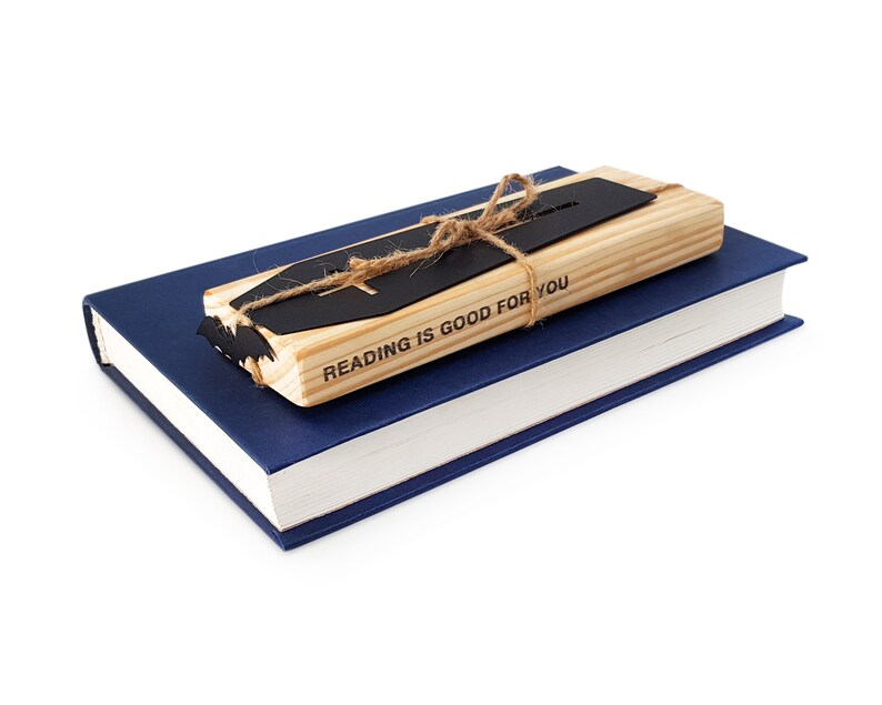 Black metal coffin-shaped bookmark with a wooden engraved block tied together with twine, resting on top of a closed blue hardcover book on a white background.