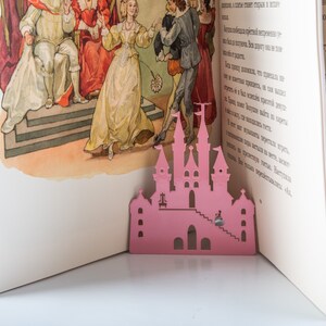 A pink metal fairy castle cutout bookmark on a book with a colorful illustration of a princess and a king in a castle