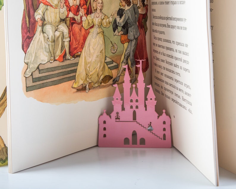 A pink metal fairy castle cutout bookmark on a book with a colorful illustration of a princess and a king in a castle