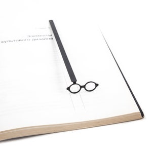 Minimal black metal page saver shaped like round architect glasses, resting on a stark design-focused book spread. Bauhaus-inspired, modernist reading accessory.