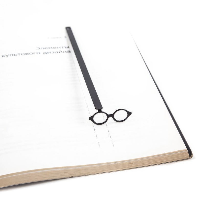Minimal black metal page saver shaped like round architect glasses, resting on a stark design-focused book spread. Bauhaus-inspired, modernist reading accessory.