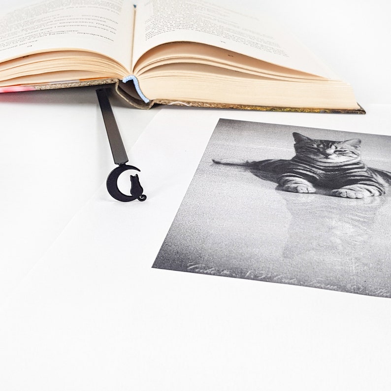 May include: A black metal bookmark shaped like a crescent moon with a cat silhouette sitting on the moon. The bookmark is laying on a white surface next to a book and a black and white photo of a cat.