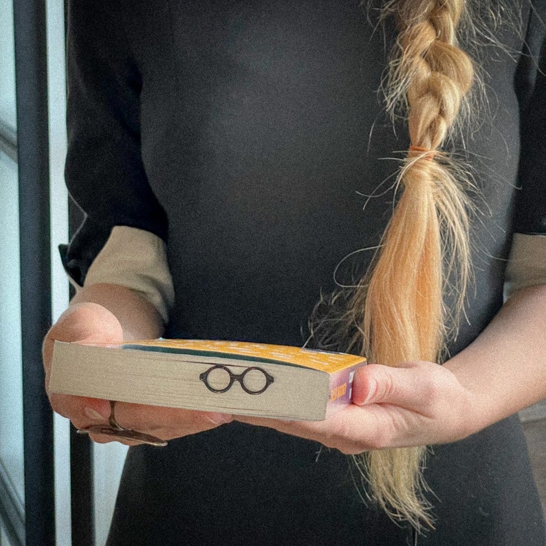 Woman holding a closed book with a black metal page saver shaped like round architect glasses peeking out. A thoughtful, design-inspired gift for modern readers.