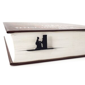 A black metal bookmark shaped like a person standing next to a building. The bookmark is placed on top of a closed book with a brown cover.