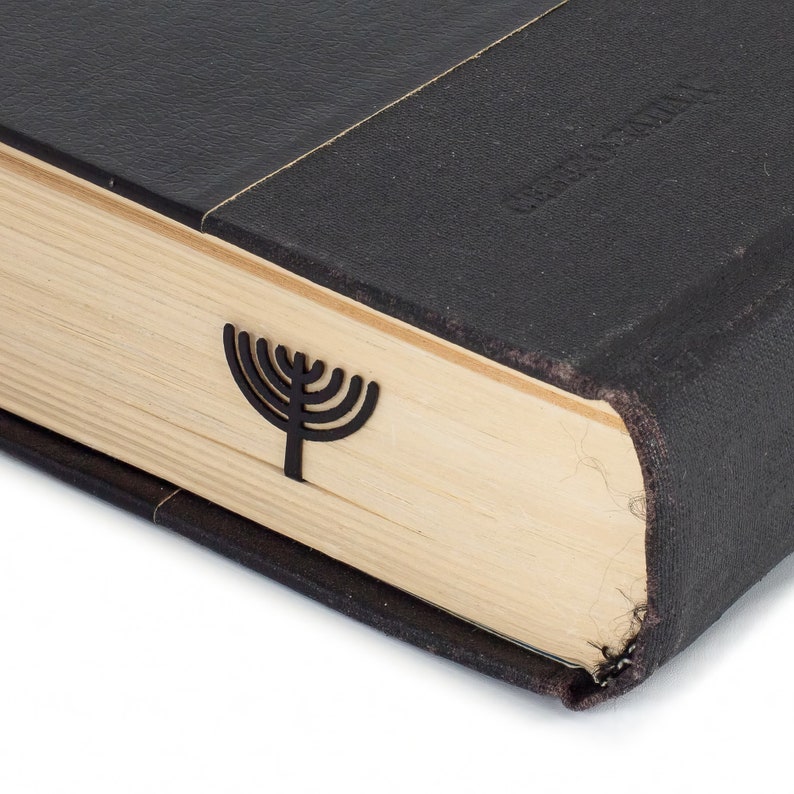 Black metal menorah-shaped page saver peeking from a closed vintage-style book. A subtle and elegant Judaica gift for book lovers