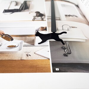 Close-up of a black metal running cat bookmark placed on an open interior design book showing images of a sink and a person pouring pet food.