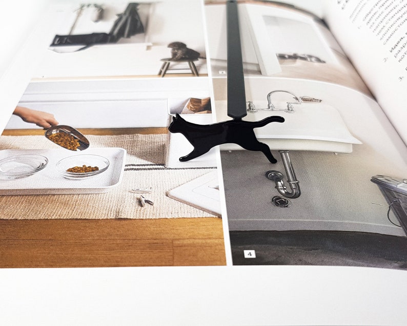 Close-up of a black metal running cat bookmark placed on an open interior design book showing images of a sink and a person pouring pet food.