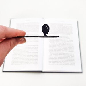 Side view of a black metal bike bookmark held between fingers, showing the thin profile and balloon detail above an open book on a white background.