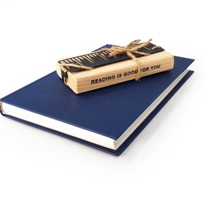 May include: A navy blue book with a light-colored spine is topped with a wooden bookmark and a black metal keyboard design. The bookmark is tied with twine and has the text "READING IS GOOD FOR YOU".