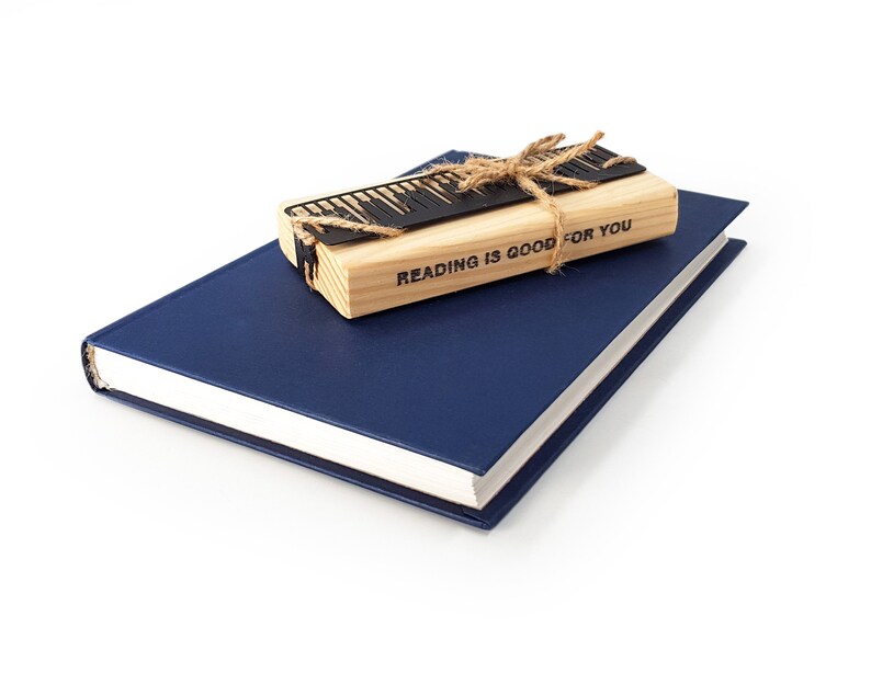 May include: A navy blue book with a light-colored spine is topped with a wooden bookmark and a black metal keyboard design. The bookmark is tied with twine and has the text "READING IS GOOD FOR YOU".