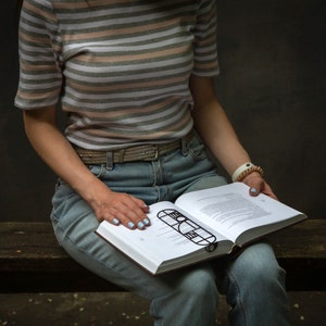 Woman sitting on a bench reading a book with a black geometric Bauhaus-style metal bookmark placed across the pages — stylish and modern reading accessory.